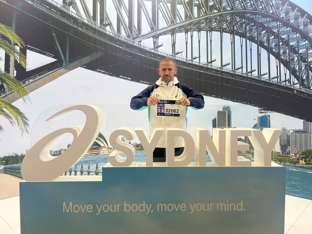 Denny holding his race bib in front of the ASICS Sydney sign, Harbour Bridge behind